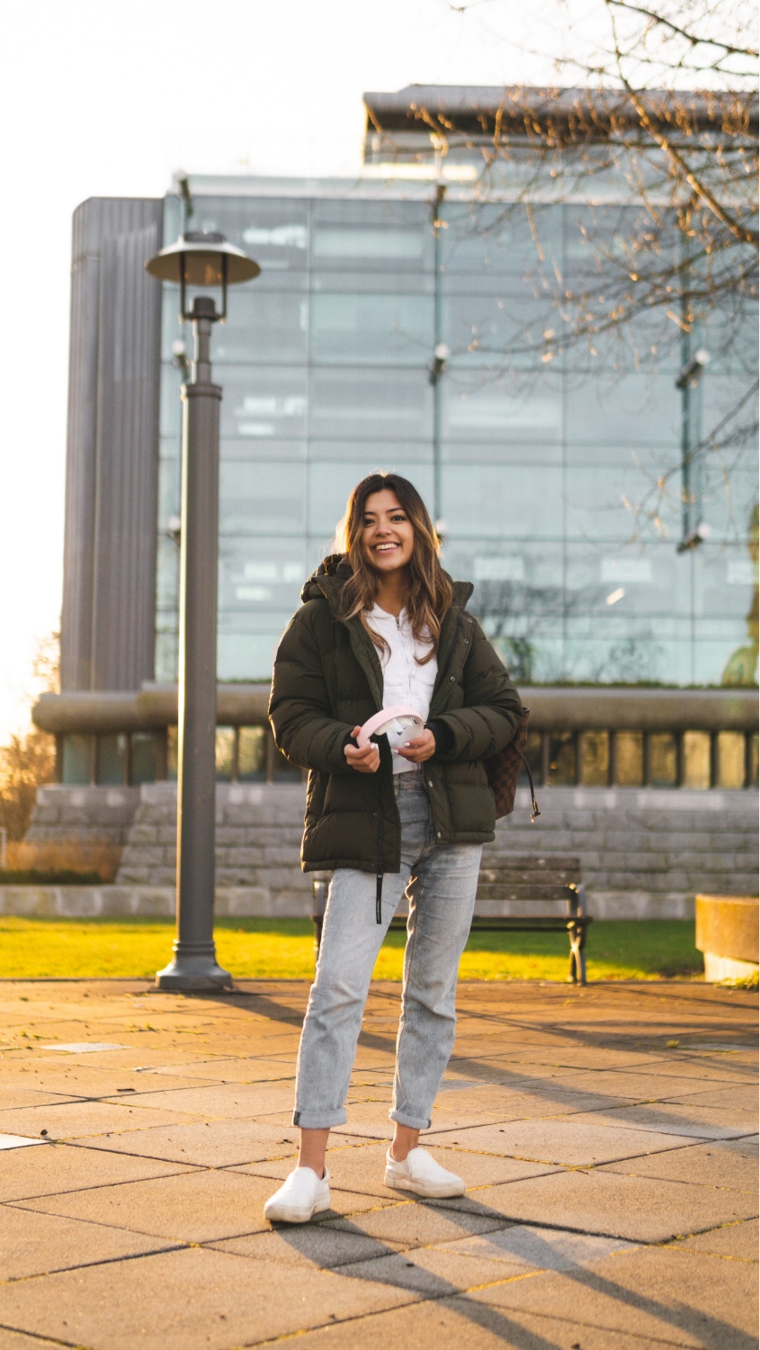 A photo of a student posing on their university campus. They are smiling at the camera and holding pink headphones.