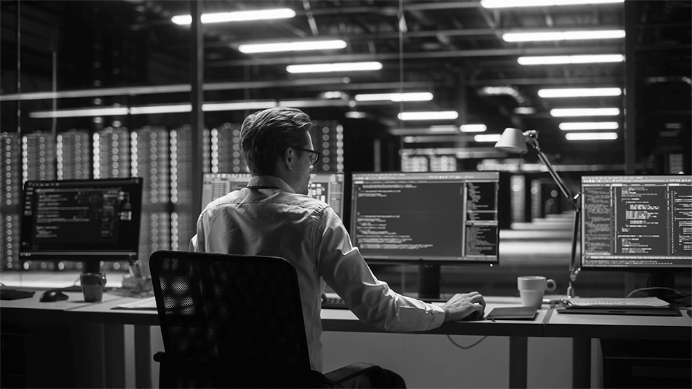 A black and white image of a man working in front of four computer screens containing code.