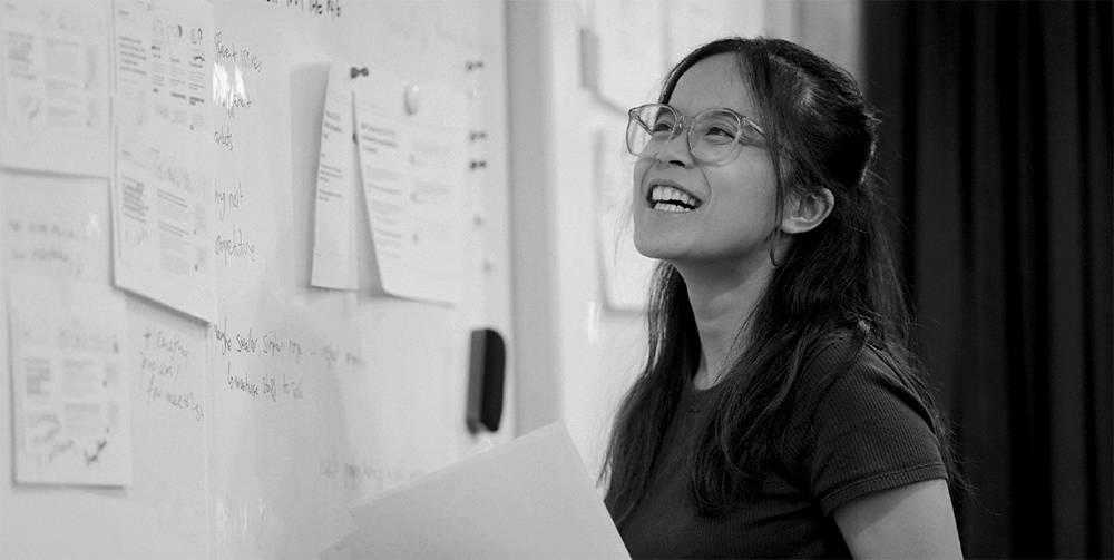 A black and white image of a teacher with glasses smiling and looking at a whiteboard. The whiteboard is covered in pieces of paper and writing.