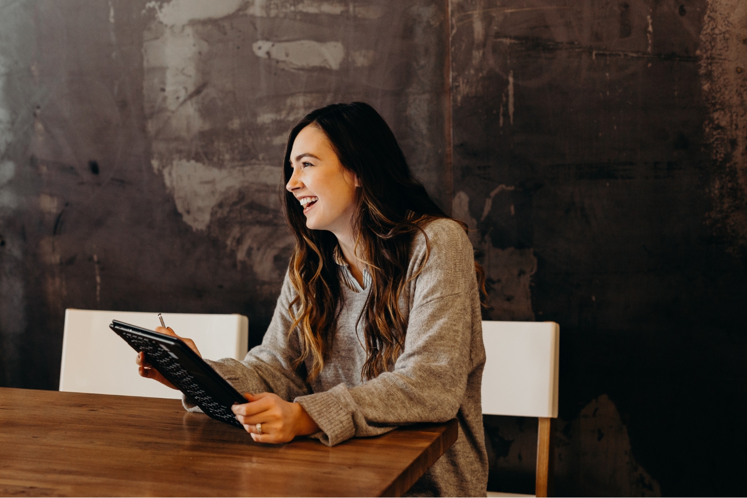 A photo of a woman sitting at a table smiling while holding a notebook and pencil.