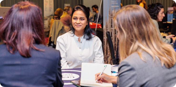 A photo of two women sitting at a table taking notes while they speak to another woman.