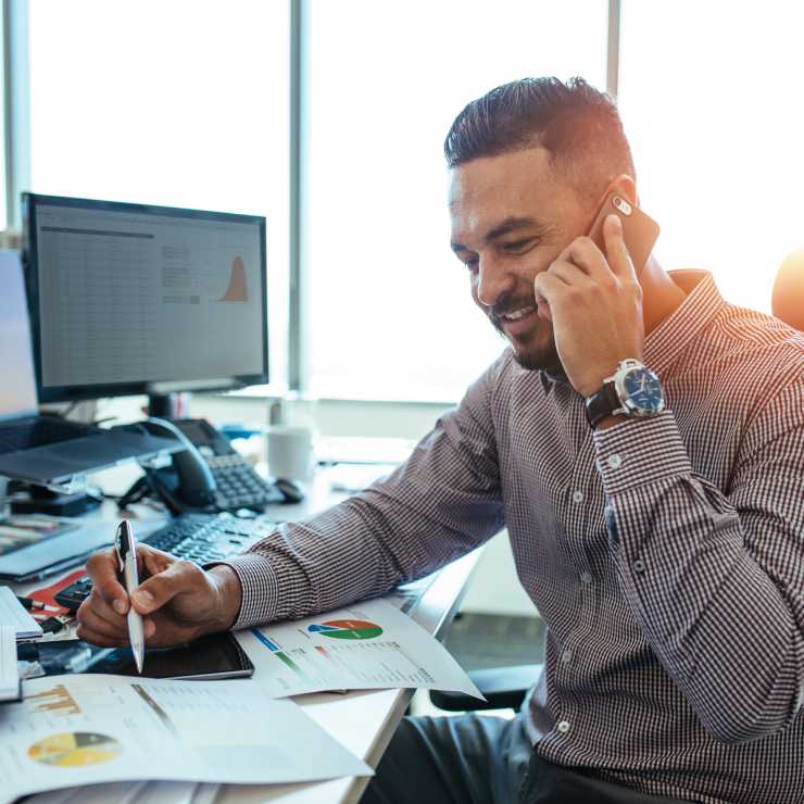 A man is taking a call whilst sitting at his desk and smiling.