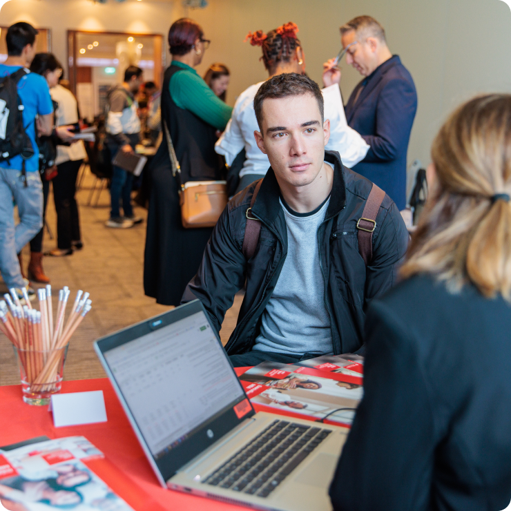 A photo of a woman sitting at a table in a crowded room with a laptop while a student sits and speaks with her.