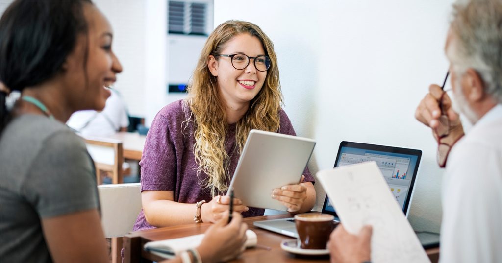 Three students smiling and laughing as they sit at a desk in front of laptops and notebooks.