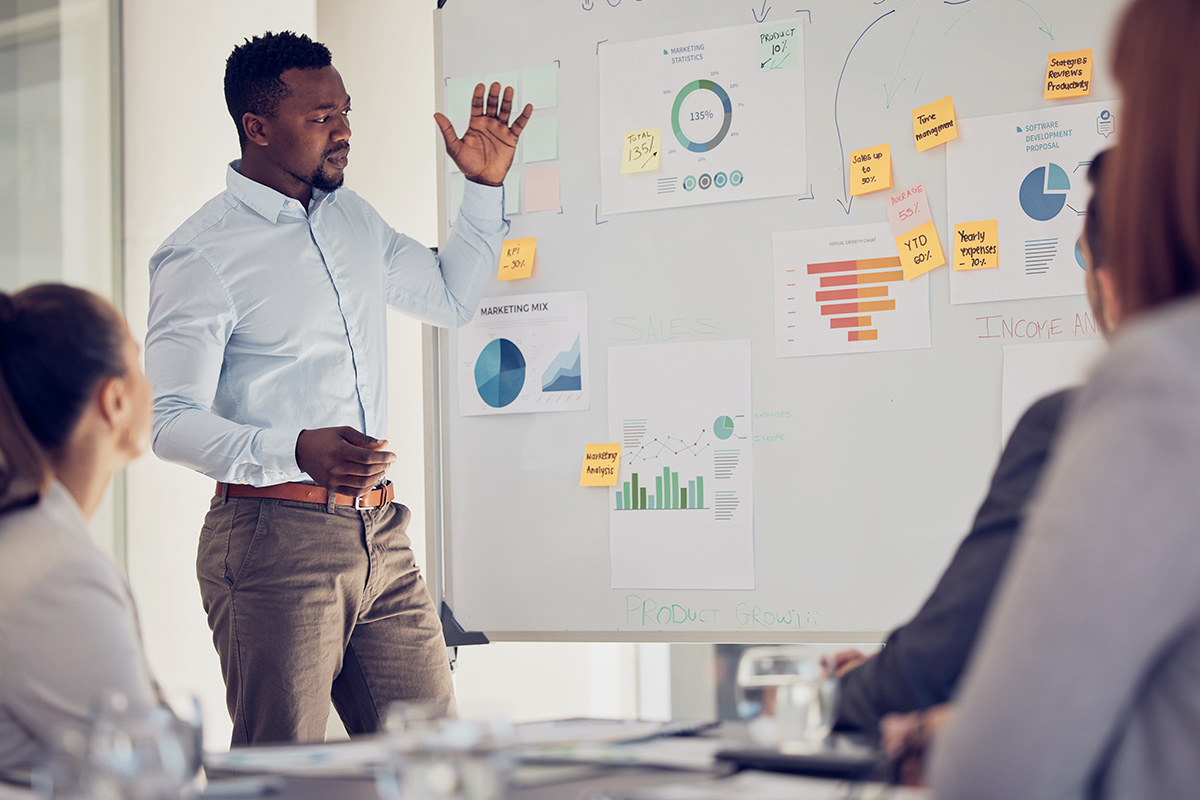 A man presents to his colleagues in front of a whiteboard covered in graphs and sticky notes