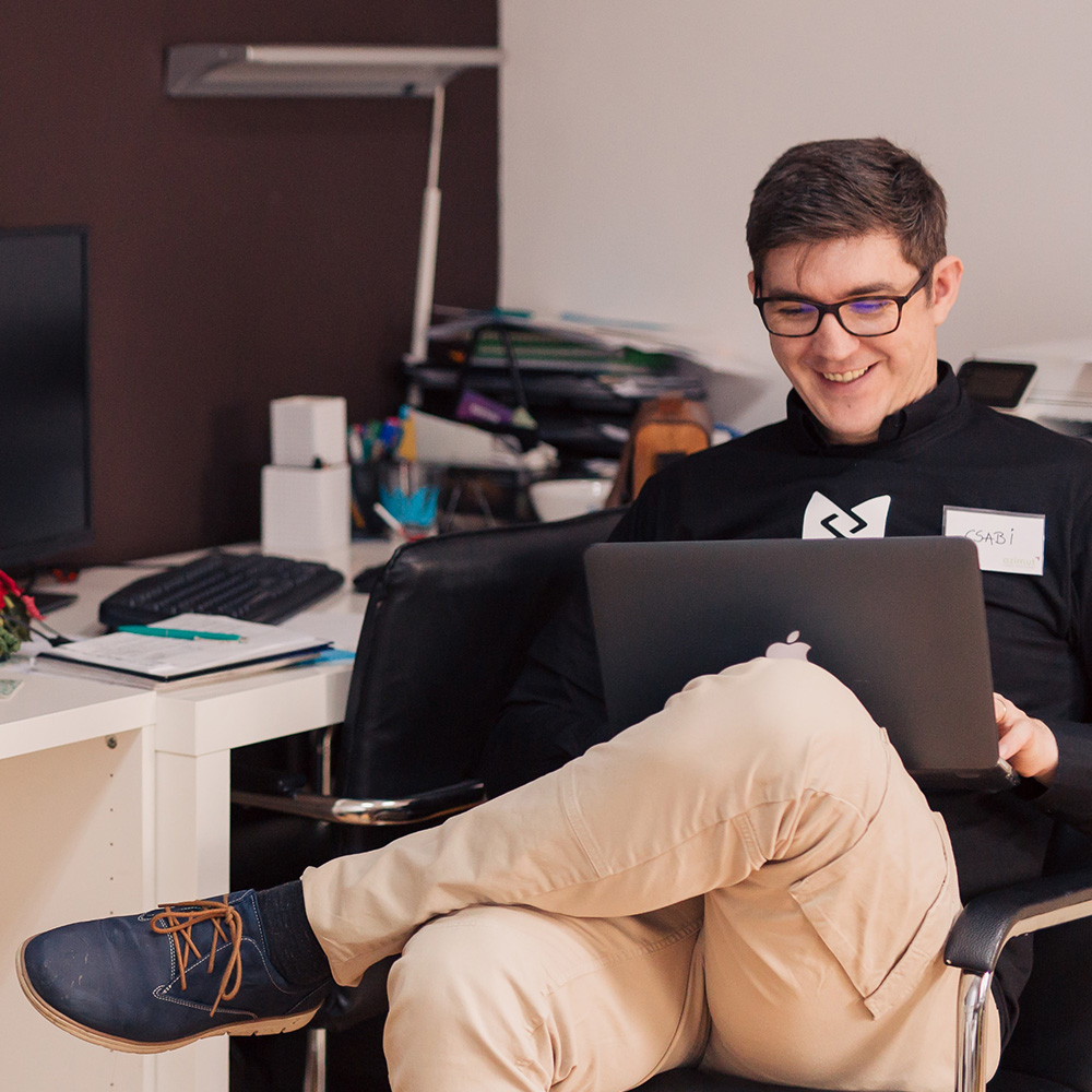 A student sitting in a chair balancing their laptop on their folded legs. The student is looking down at the laptop and smiling. He is wearing a name tag which reads ‘CSABI’.