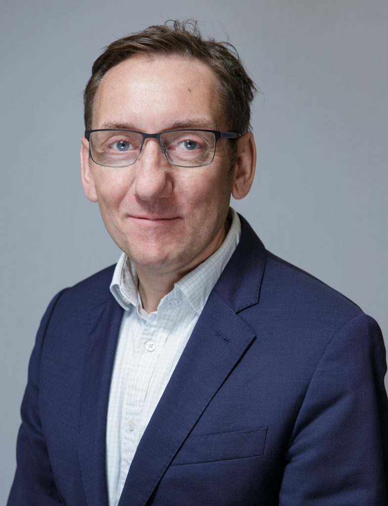 A portrait photo of Edward Harcourt, Senior Vice President of Enrolment Solutions, wearing a suit and glasses. Mr Harcourt is smiling whilst looking at the camera.