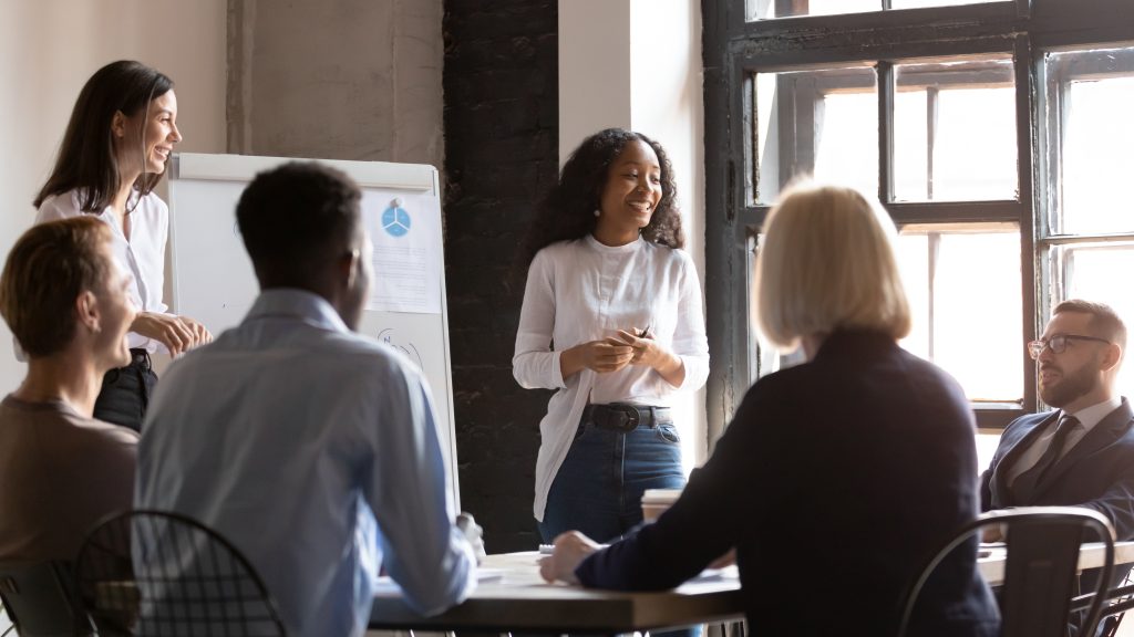 Two women stand up presenting to a group sitting at a table. There is a whiteboard with a chart on it behind them.