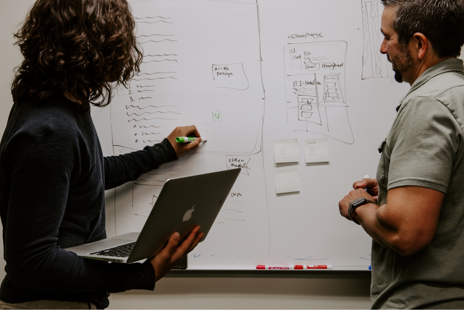 Two colleagues work on a large whiteboard. One of them watches as the other writes while holding a laptop.