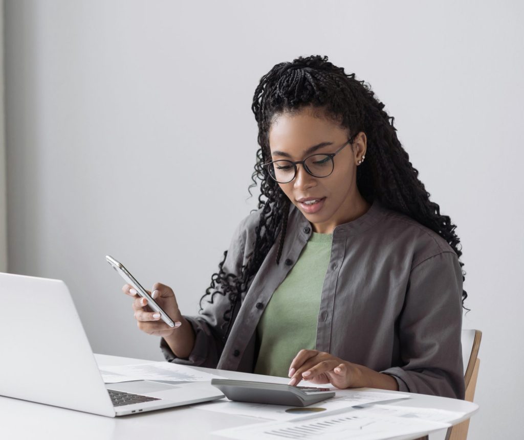 A woman in glasses works at a desk covered with paper and a laptop. She’s holding a phone and typing on a calculator.