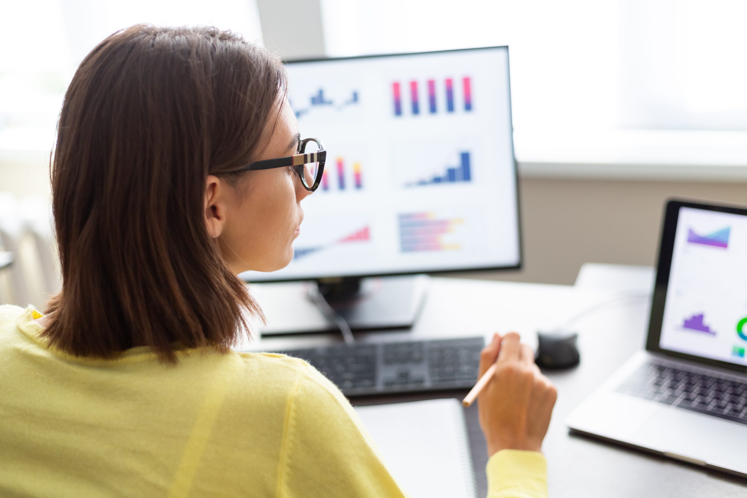 A woman in glasses works at a desk with a laptop and monitor showing various graphs and charts.