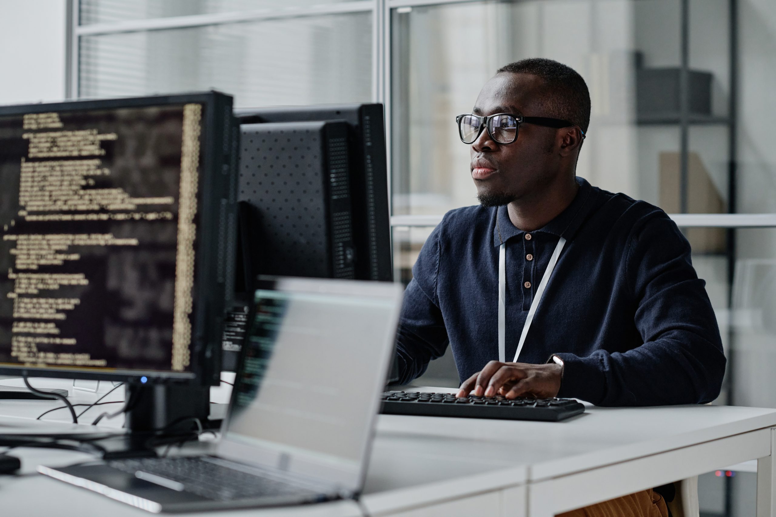 An employee with glasses works at a computer. The screen in front of him is full of code.