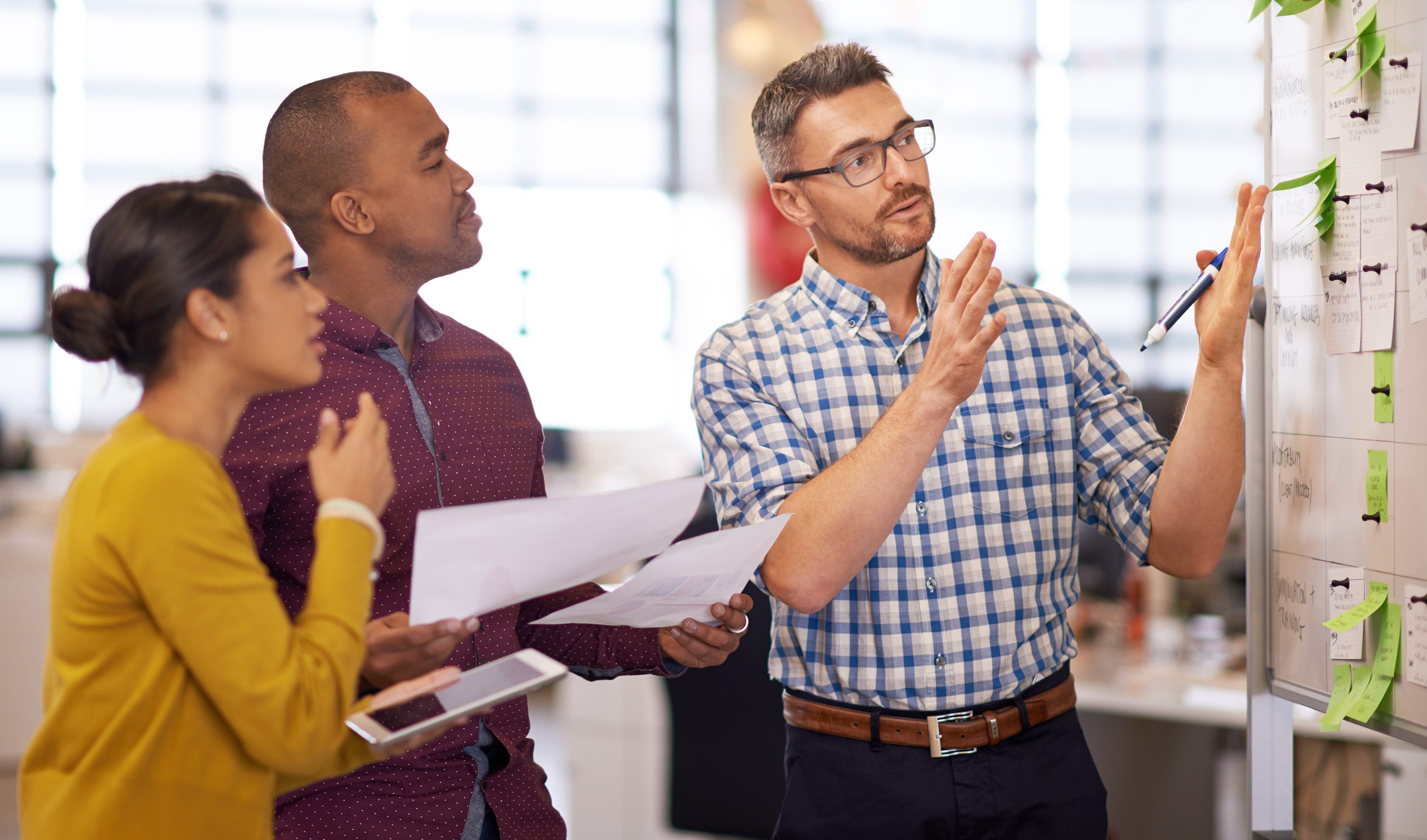 Three employees are having a conversation whilst gathered around a whiteboard covered in sticky notes.