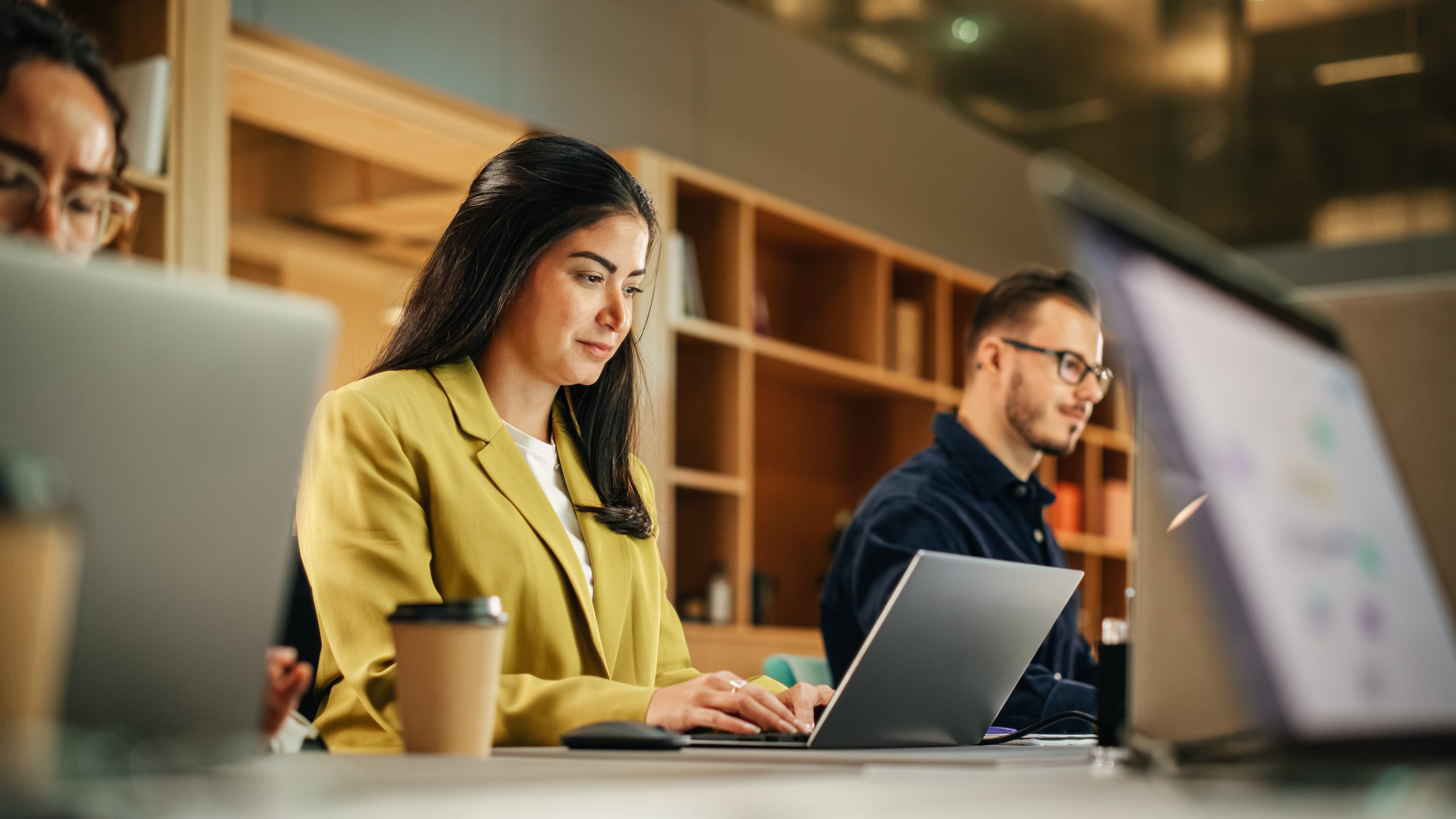 A woman in a yellow blazer works on a laptop surrounded by other working colleagues.