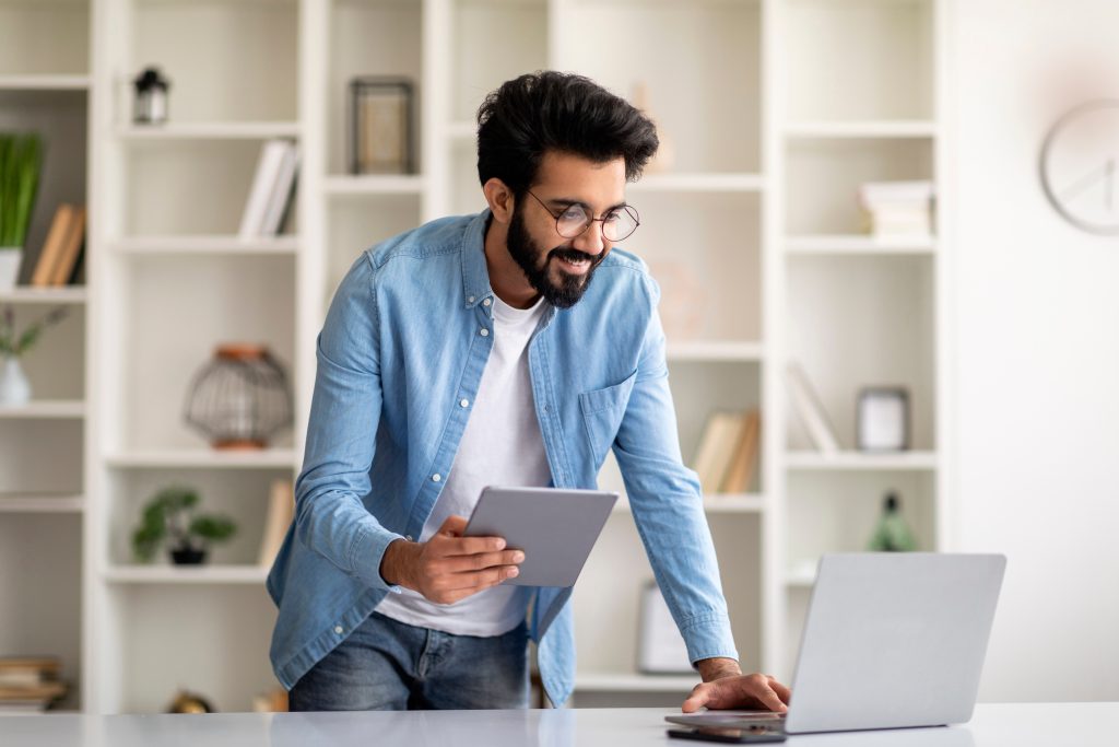 A man in glasses stands over a desk working on a laptop and tablet.