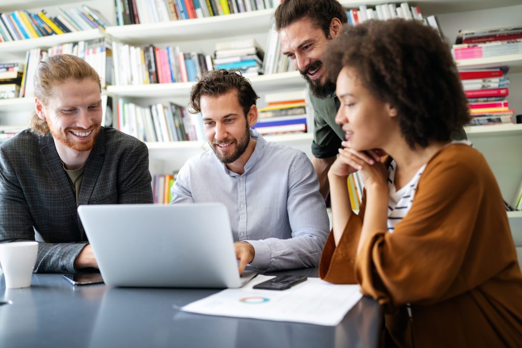 Four colleagues crowd around a laptop, smiling and conversing.