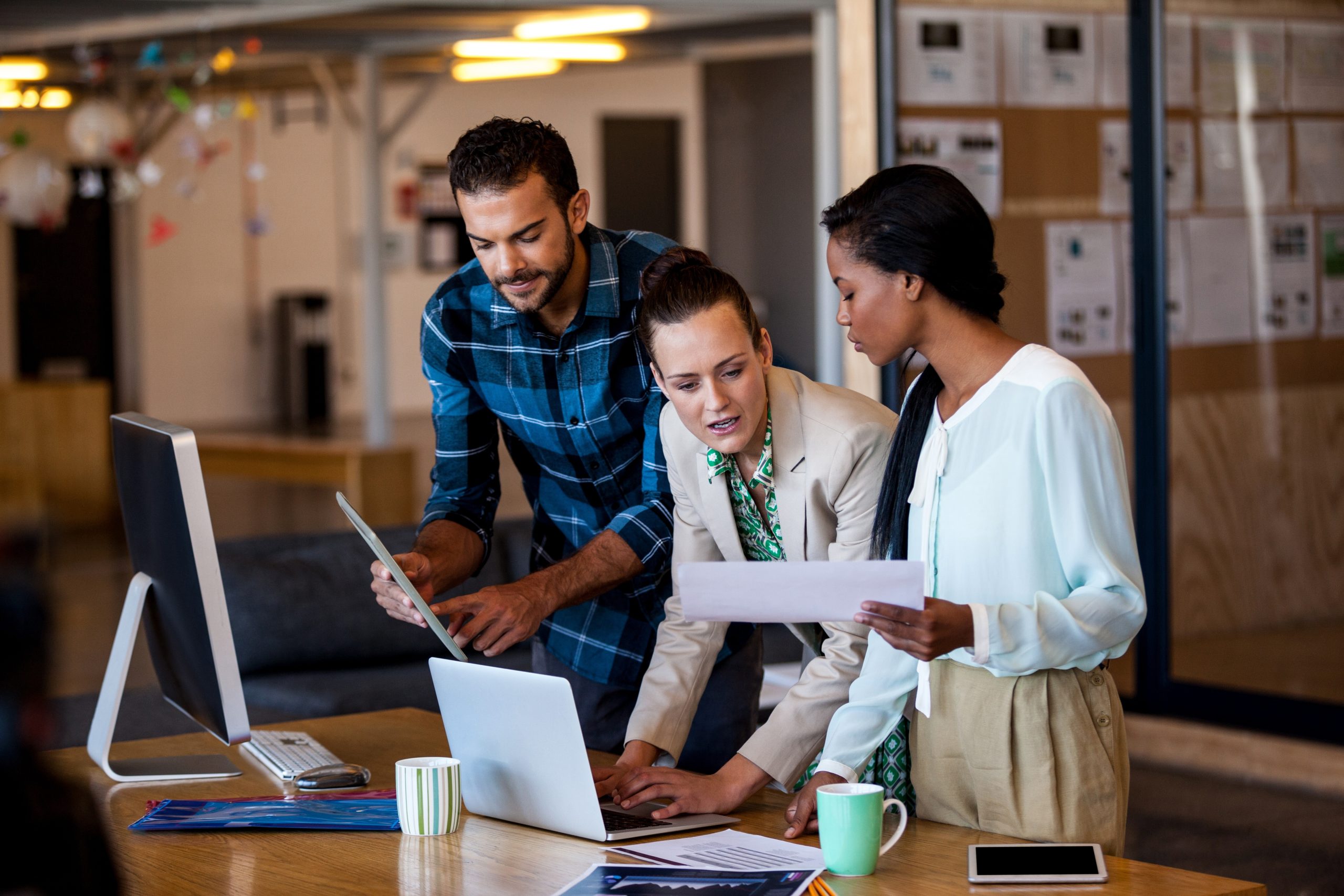 Three employees are gathered around a desk working together and comparing notes