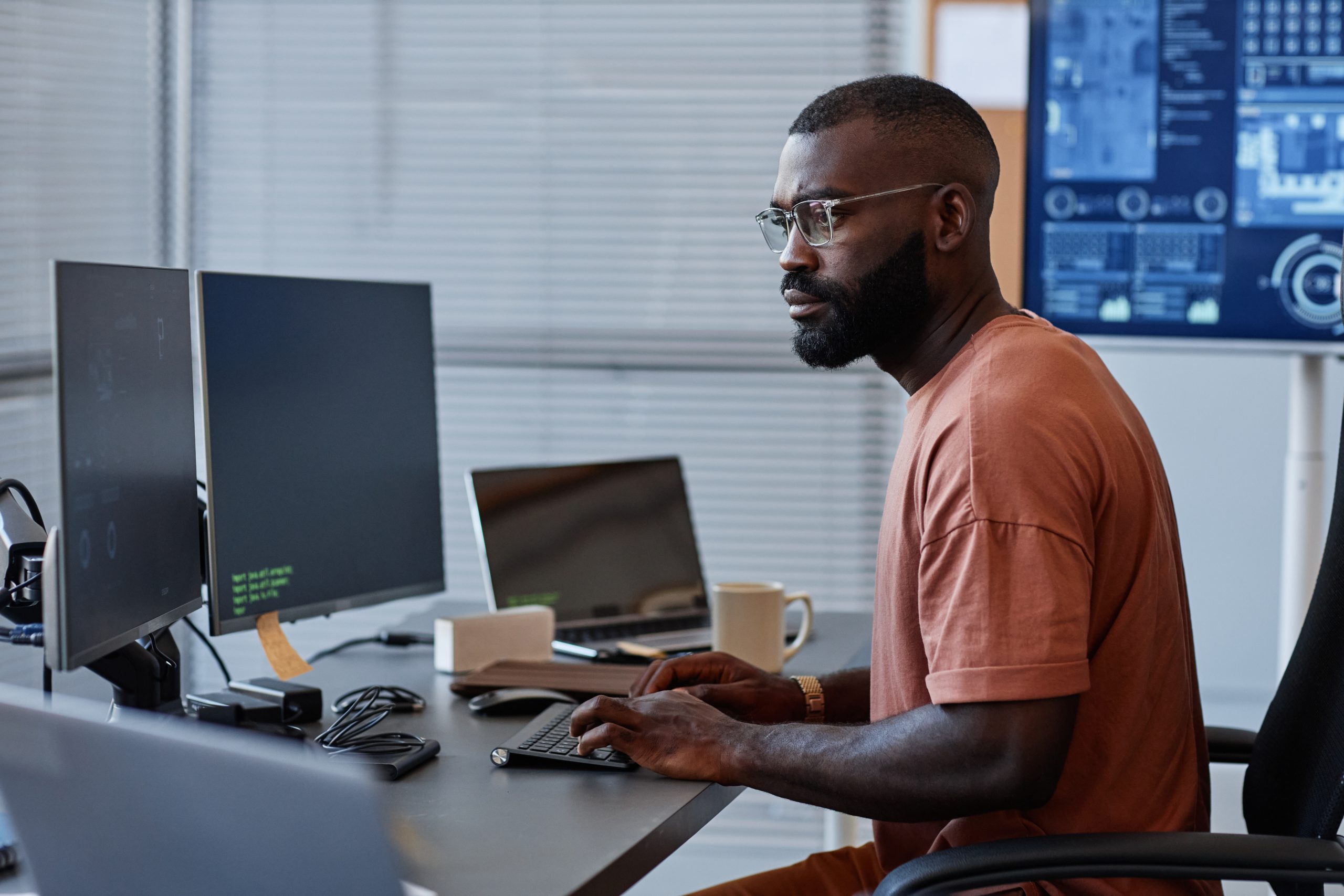 A man is sitting at a desk writing code. He is using two monitors and a laptop.