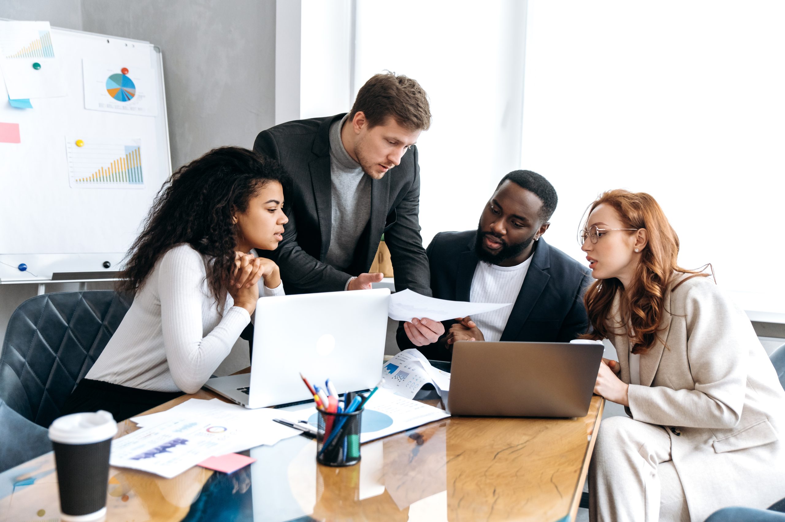 Four employees are gathered around a table in a meeting room. There are two laptops on the table. Three of the employees are sitting down and the fourth employee is standing over them whilst holding a piece of paper.