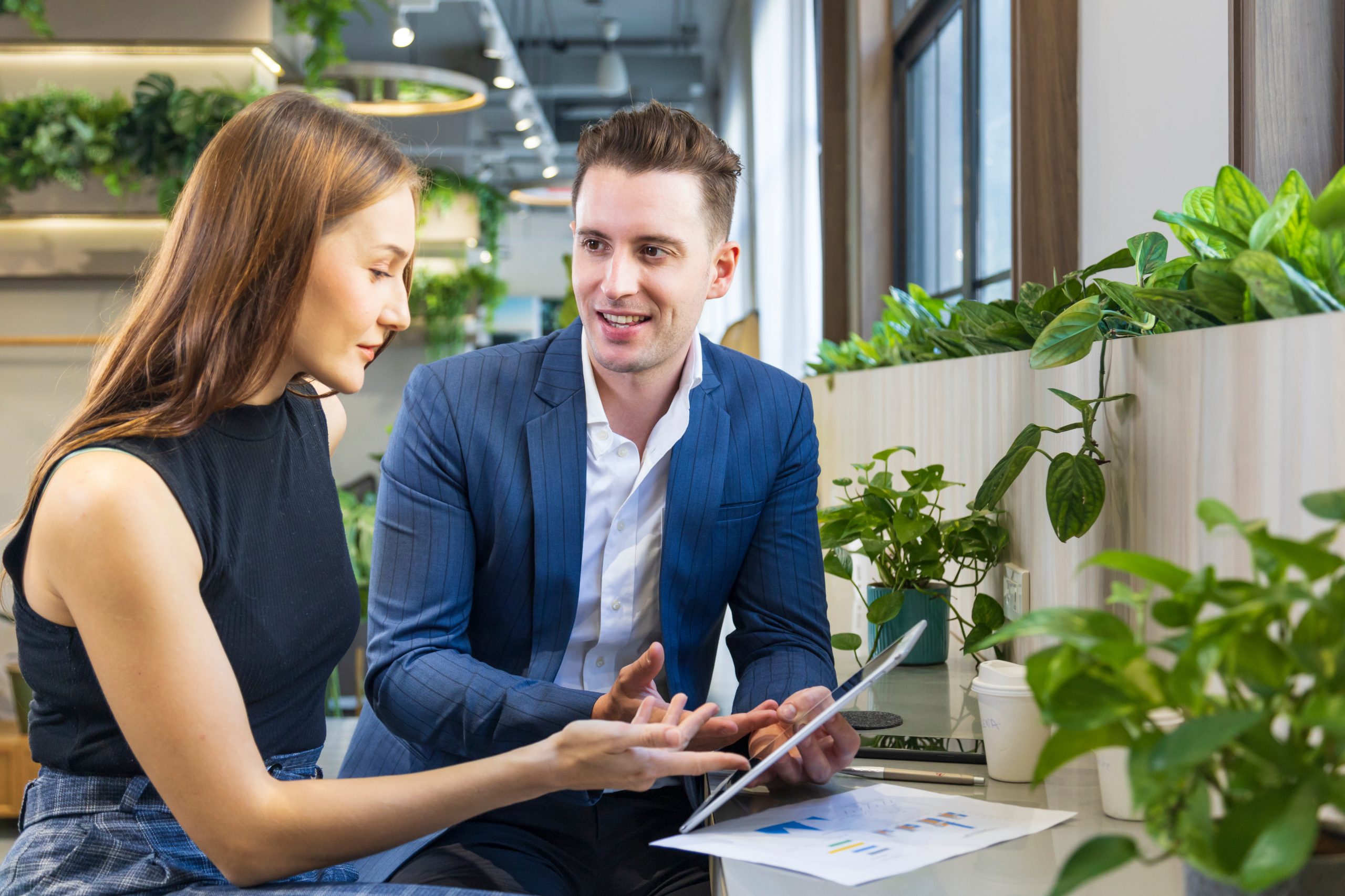 Two employees are sitting down at a desk and having a conversation whilst looking at a tablet.