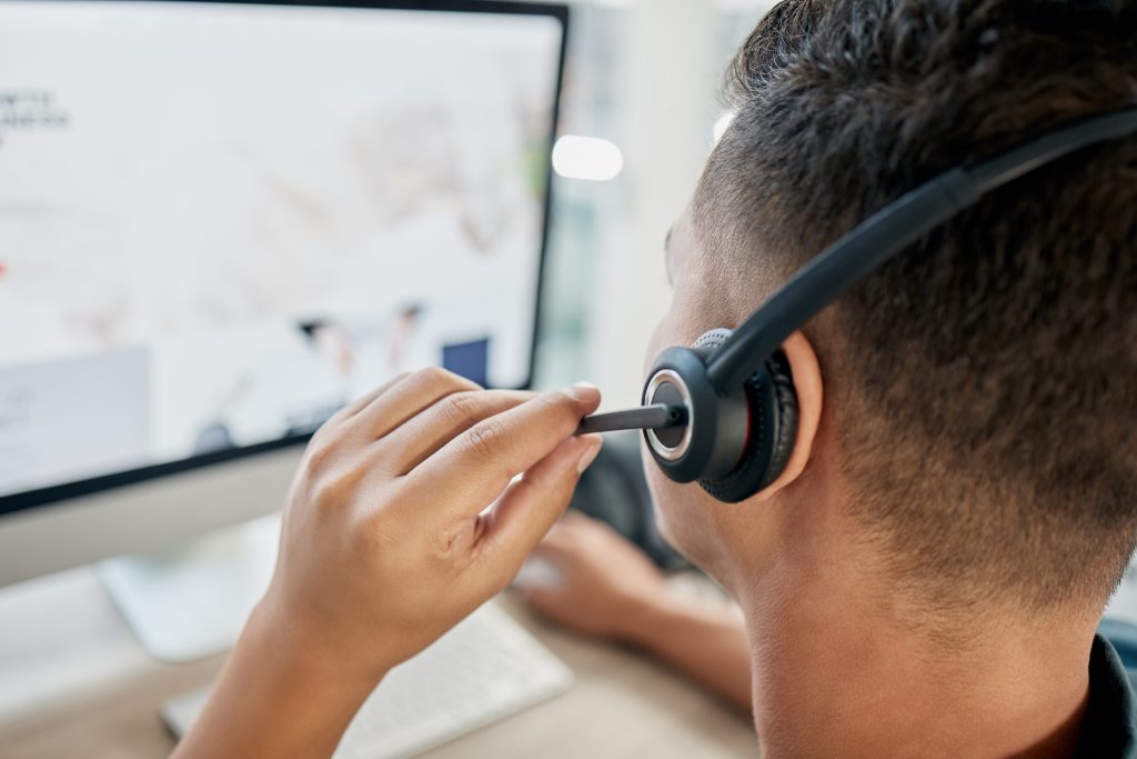 A photo of the back of a person’s head. The person is sitting at an office desk and wearing a headset.