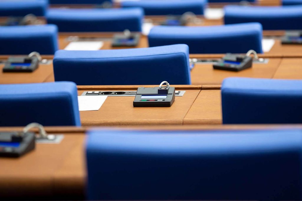 A view across a large meeting room/lecture theatre. The seats are blue and the desks wooden with built-in audio equipment.